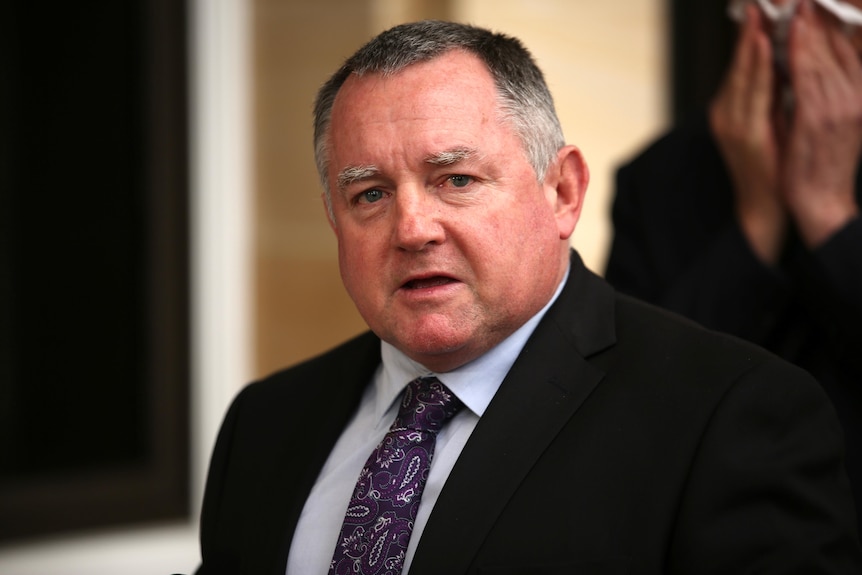 A man with greying hair and a black suit speaks at a press conference