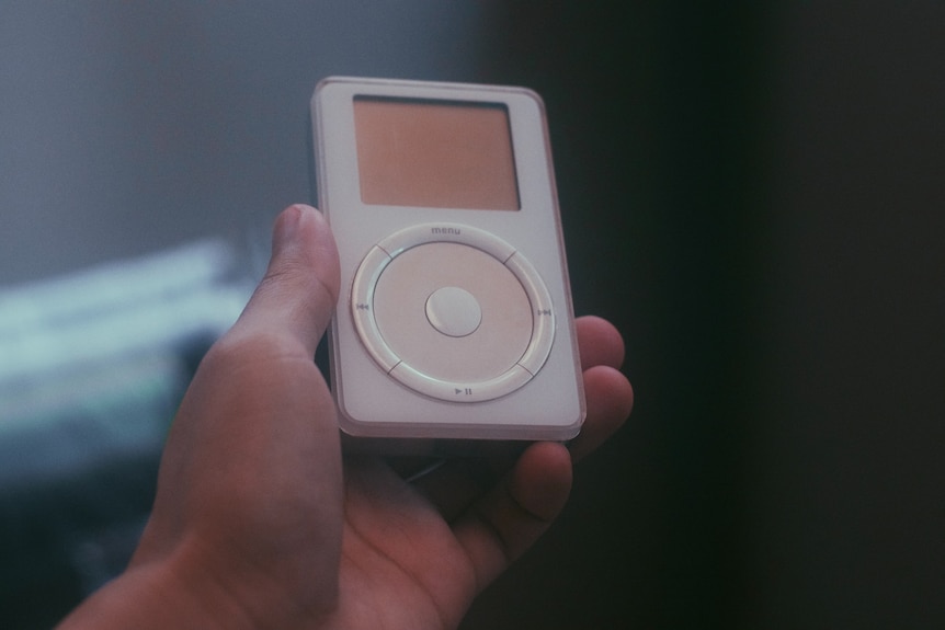 A hand holds a first generation Apple iPod, against a dark background