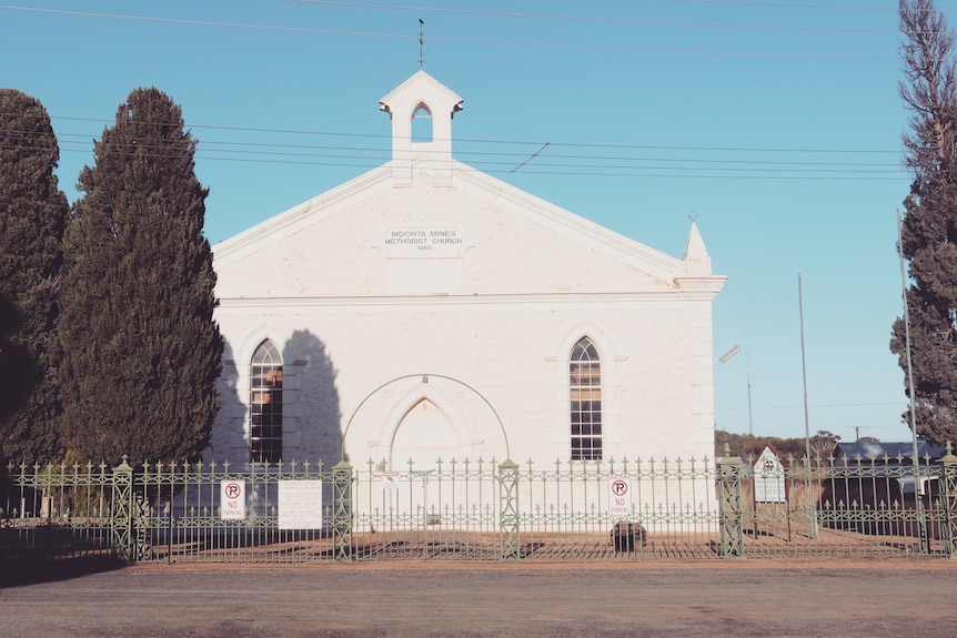 A historic white church behind an iron fence under a blue sky, with two tall ornamental bushes in front.