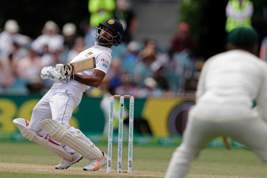 A batsman sways backwards with his bat at his waist, looking behind him towards a crouching figure in the foreground