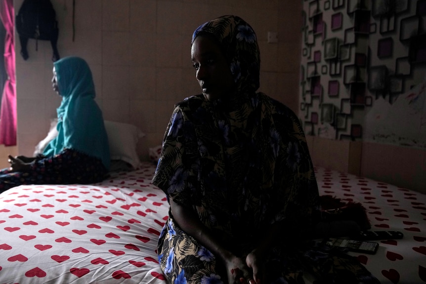 Two Sudanese women sit on the edge of a bed.