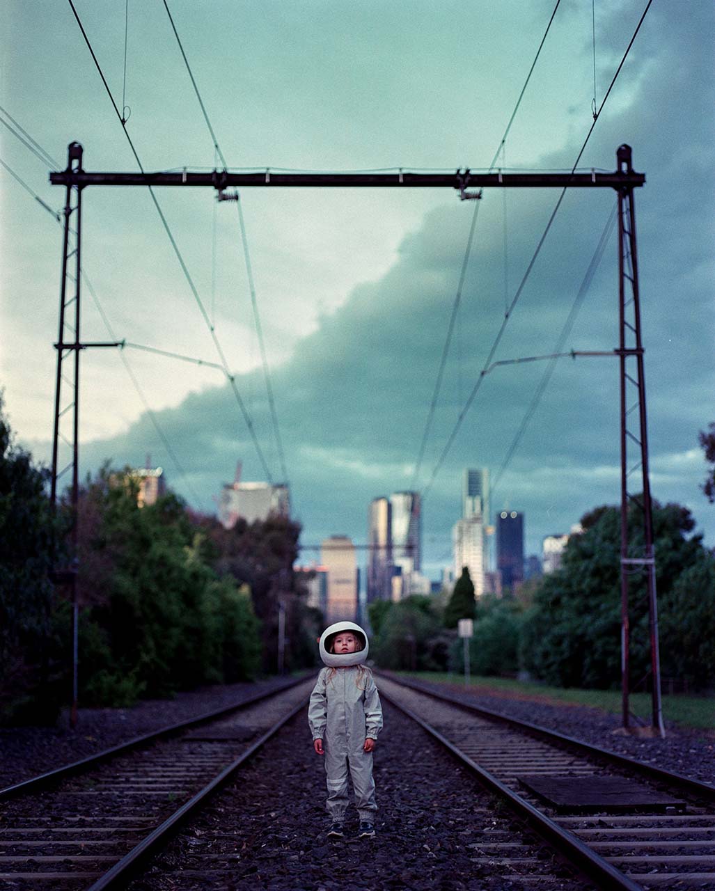 A young girl wearing a spacesuit costume and helmet stands on a train track with a city skyline behind her