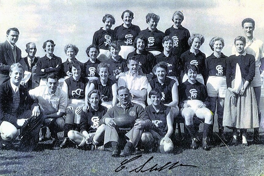 A group of young women in Carlton football jumpers sitting around a man holding a football.