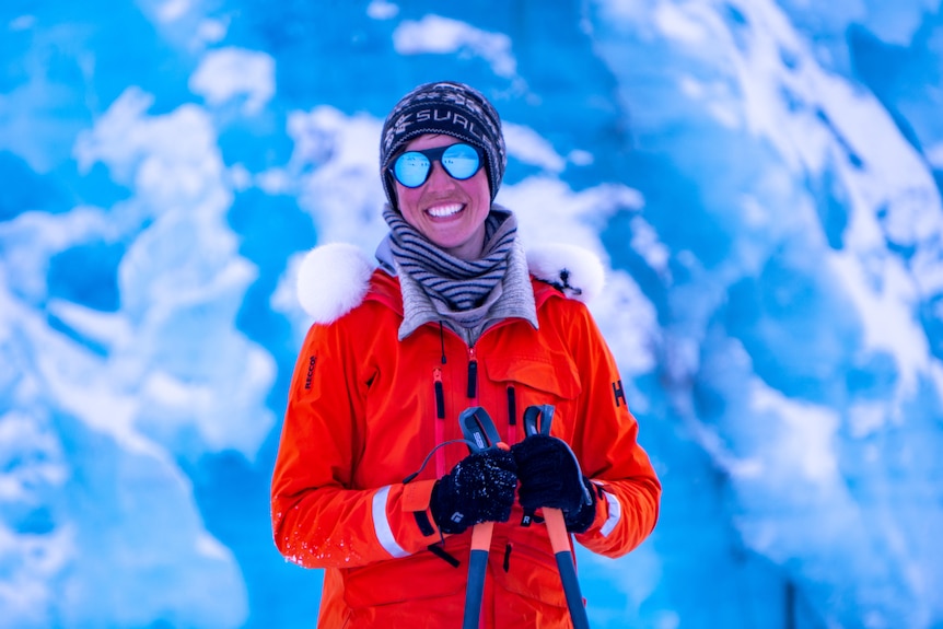 Portrait of Heidi in a beanie, gloves and a big red jacket in front of a glacier.