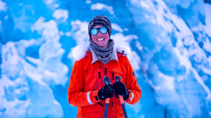 Portrait of Heidi in a beanie, gloves and a big red jacket in front of glacier.