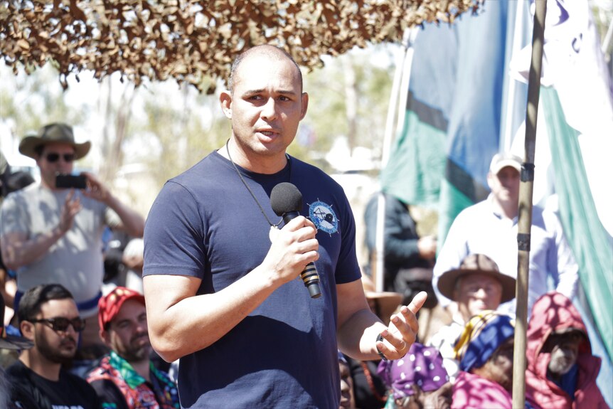 A man in a blue T-shirt standing up and addressing a crowd of people in a bush environment, on a sunny day.