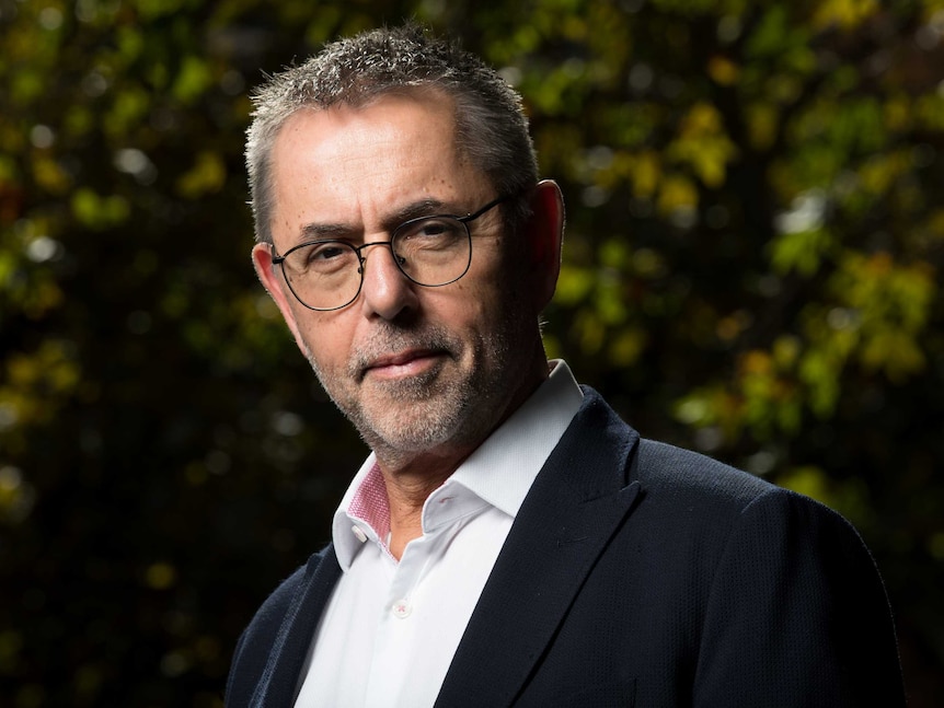 A man in glasses stares down the camera with a serious look on his face, he is surrounded by trees