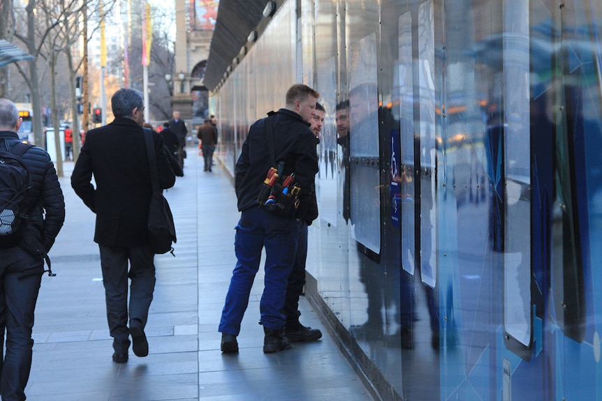 Two men in work gear look through a plastic window in a high black wall.