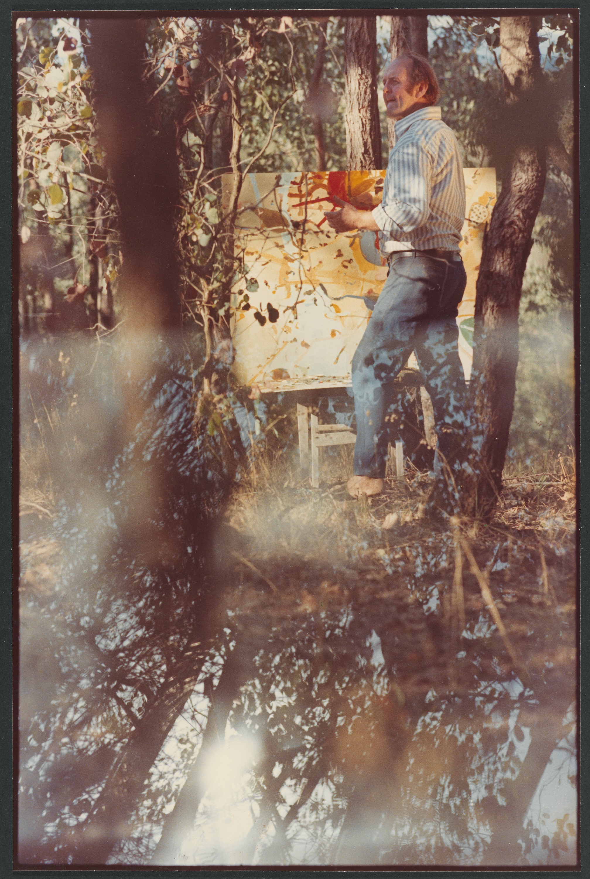 A photograph of a middle-aged man standing at an easel, painting in the Australian bush