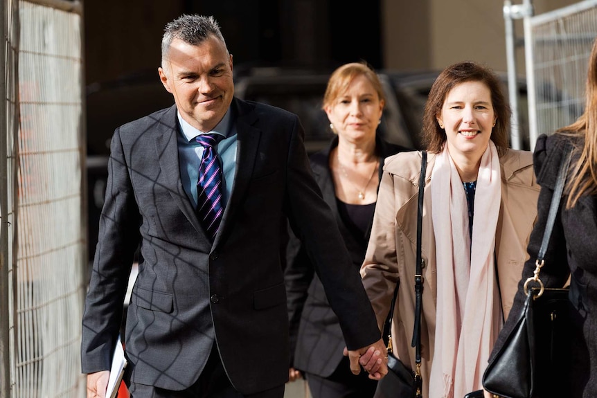 Stephen Kaless grinning as he walks outside court wearing a suit and tie and holding his wife's hand, with a woman behind them.