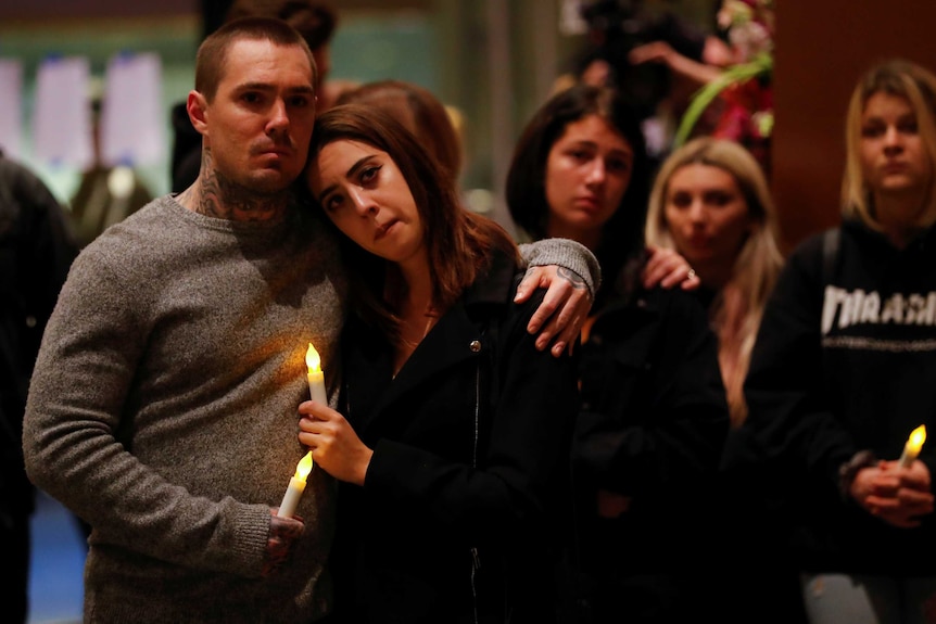 A couple stand in the foreground hugging each other and holding a candle with several other people gathered in the background