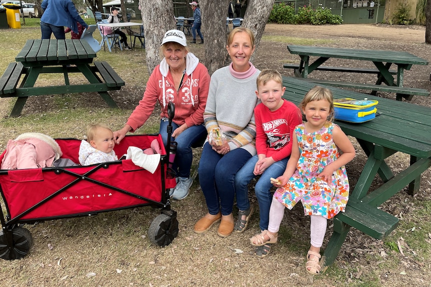 Two women, two children and a baby sitting on a picnic table smiling at the camera.