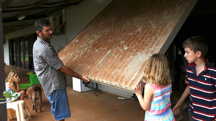 A man in a checked shirt and two children pull down a large cyclone shutter over a window on the porch of their homestead.