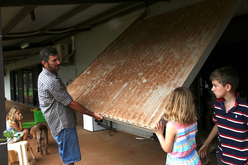 A man in a checked shirt and two children pull down a large cyclone shutter over a window on the porch of their homestead.