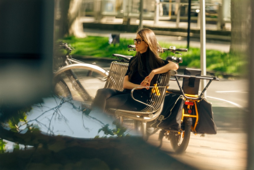 A woman with long hair and a black top and jeans sits on a metal bench next to a yellow bike