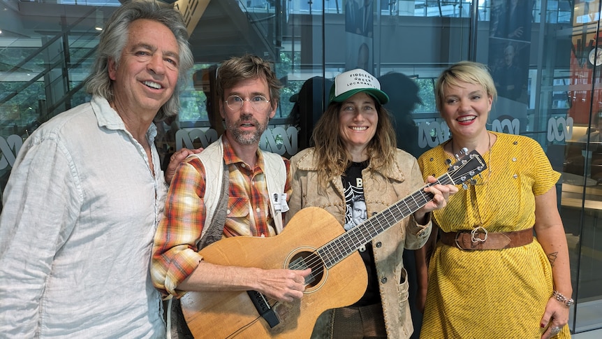 Willie Watson and Melissa Carper smile for the camera while holding a guitar, standing between the Friday Revue hosts.