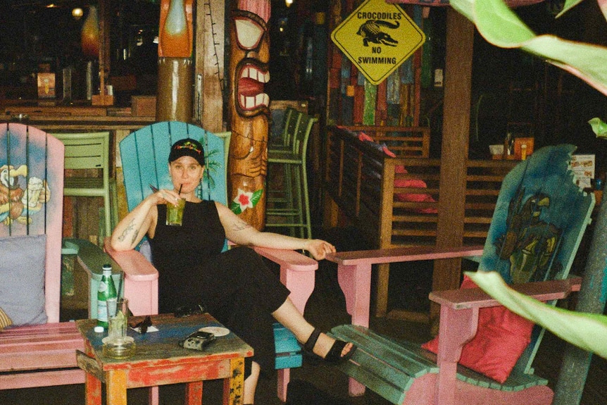 a woman wearing black singlet, pants, shoes and cap is sitting in a green and pink wooden chair sipping on a green drink. 