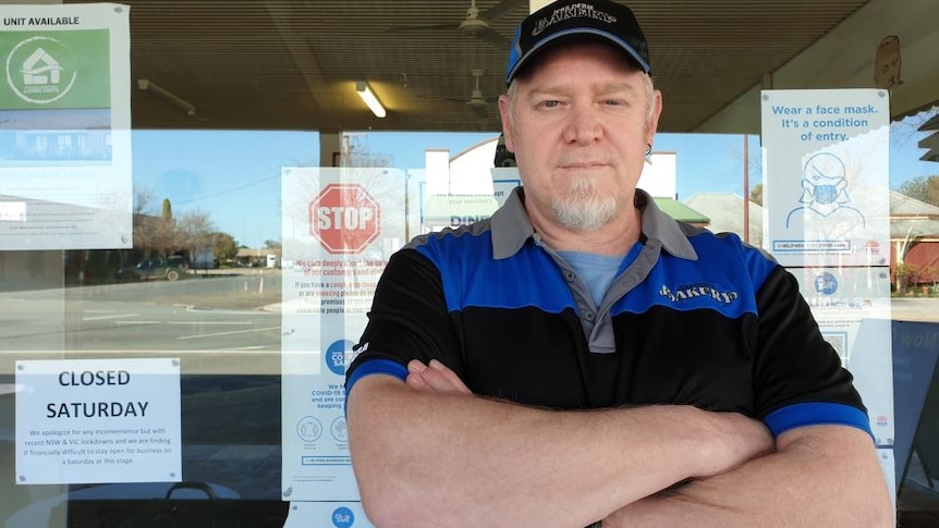 A man in a cap and a dark shirt stands in front of a bakery with his arms crossed.