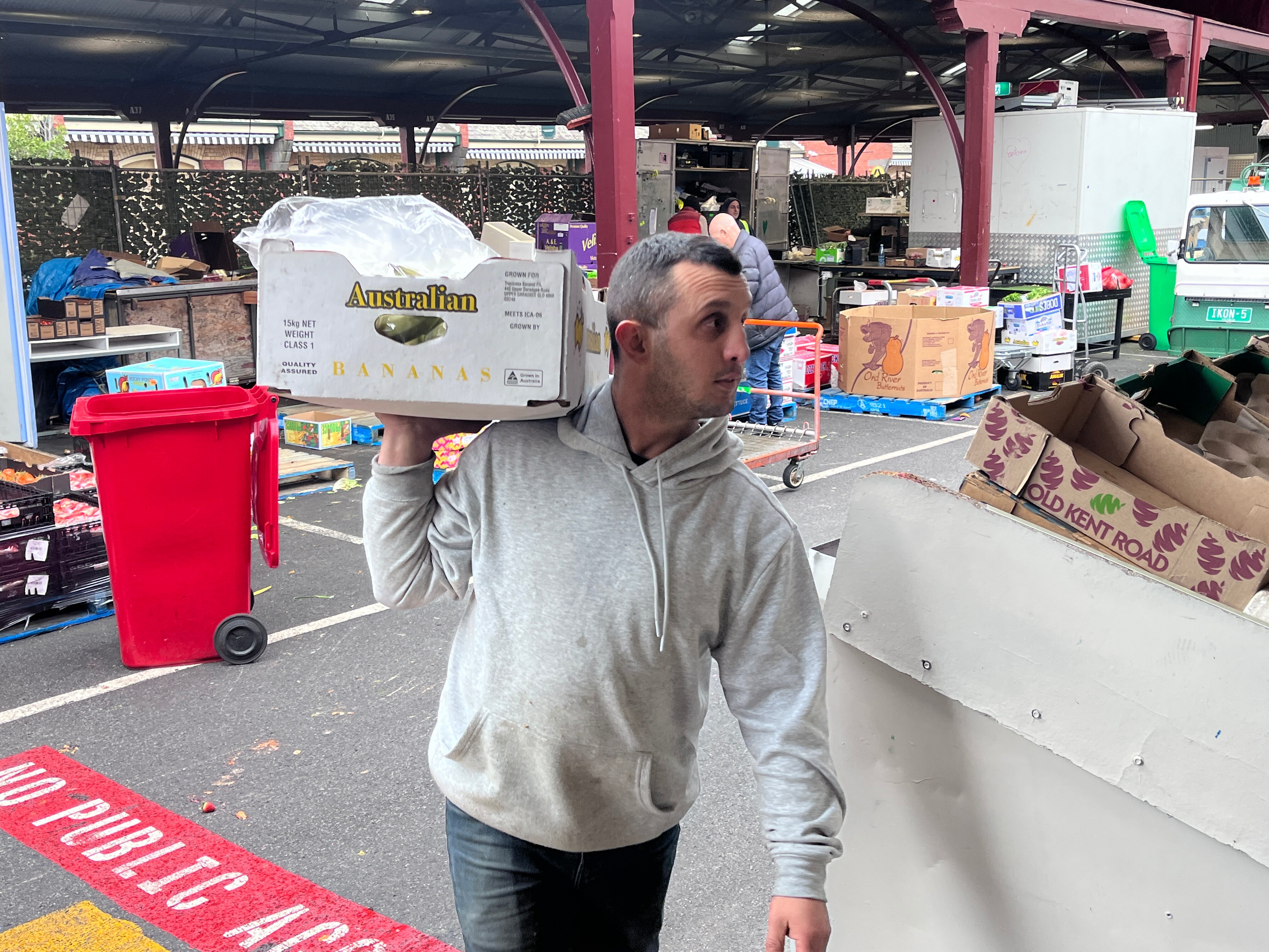 Man in a grey hoodie holding a box of fresh produce walking through a market.