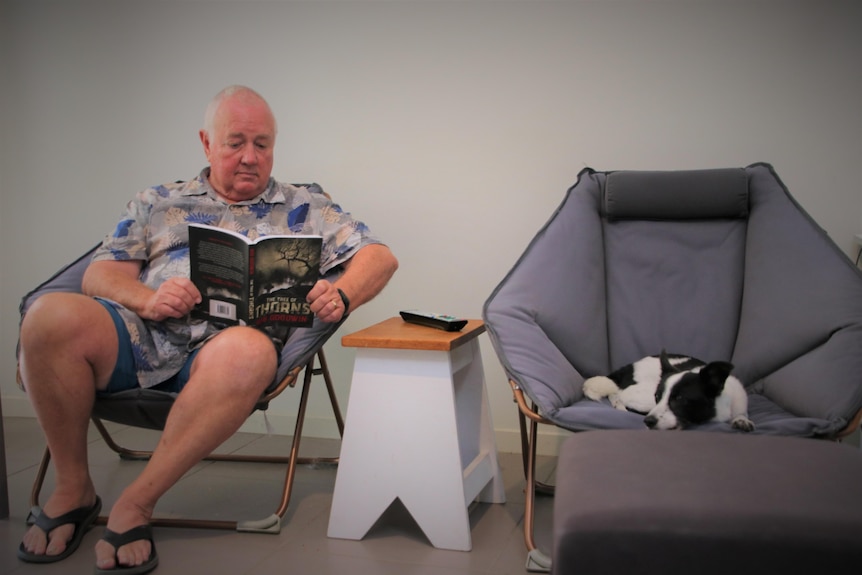 A man reading a book on a camp chair next to a dog on a camp chair.