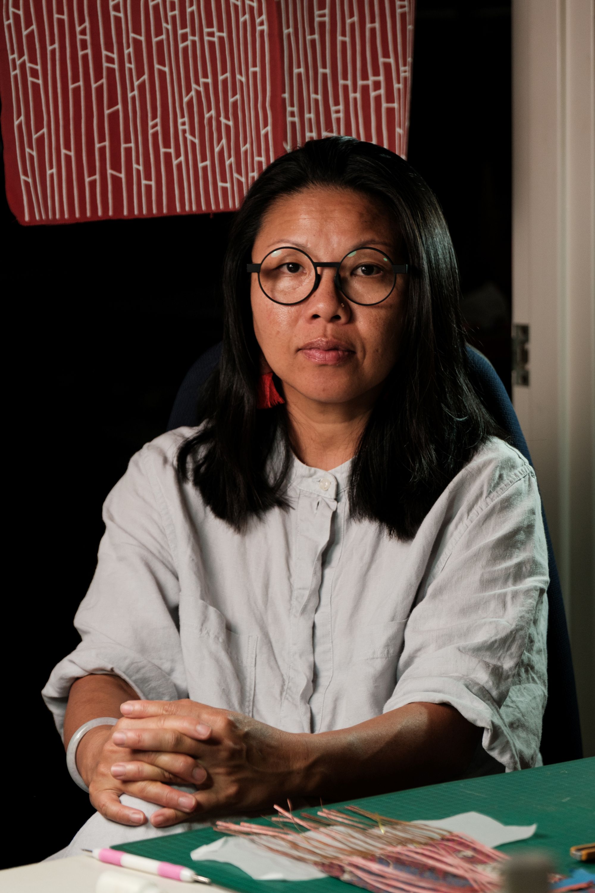 A woman with long black hair and black-framed glasses, wearing a white shirt, sitting with her hands clasped.