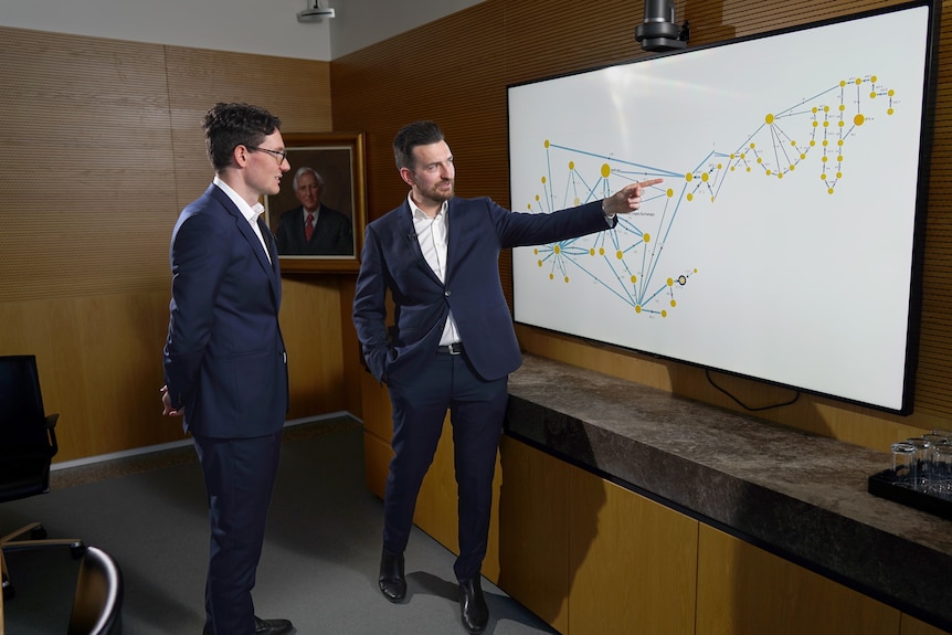 A man points at a graph on a screen while another man watches.