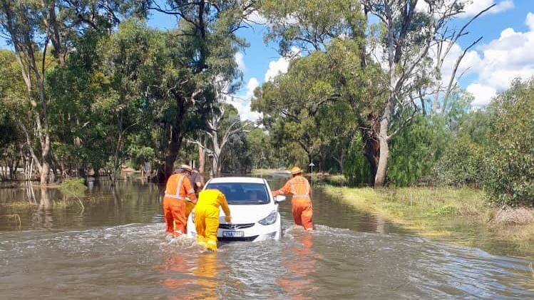 Three SES volunteers in orange jumpsuits push a car along a completely flooded road.