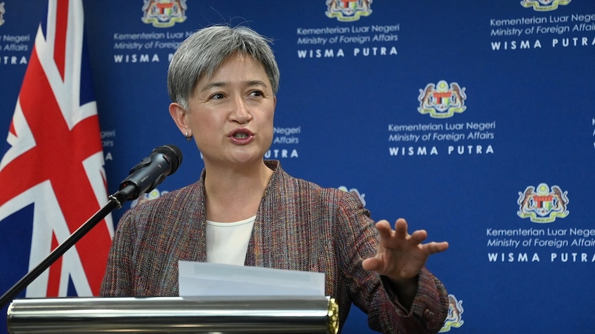 Penny Wong speaking at a microphone in front of a blue background in a press conference in Malaysia
