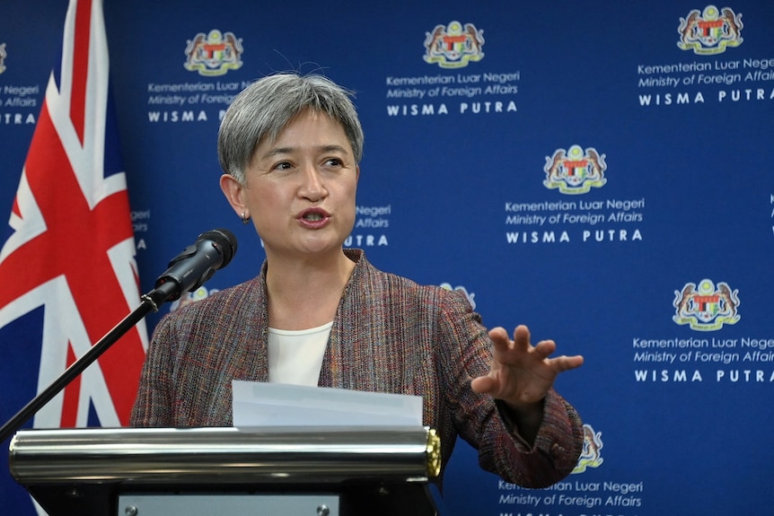 Penny Wong speaks at a microphone in front of a blue background during a press conference in Malaysia