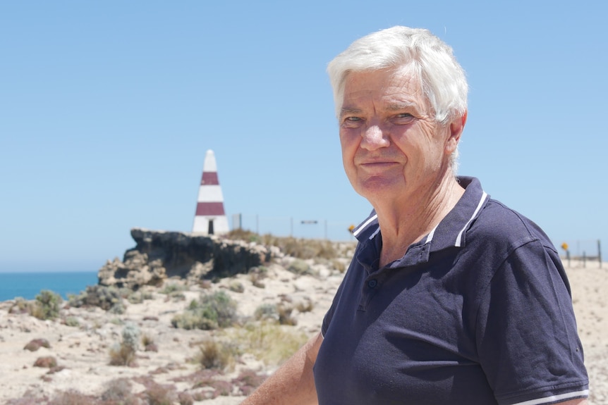 A man looks at the camera with a cliff and a red and white obelisk in the background