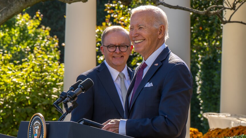 Anthony Albanese looks at Joe Biden, who is standing behind a podium in a garden. Both men are smiling.