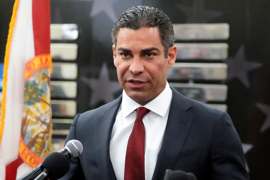 A middle-aged Hispanic man in a suit speaks into a microphone in front of a star-filled banner.