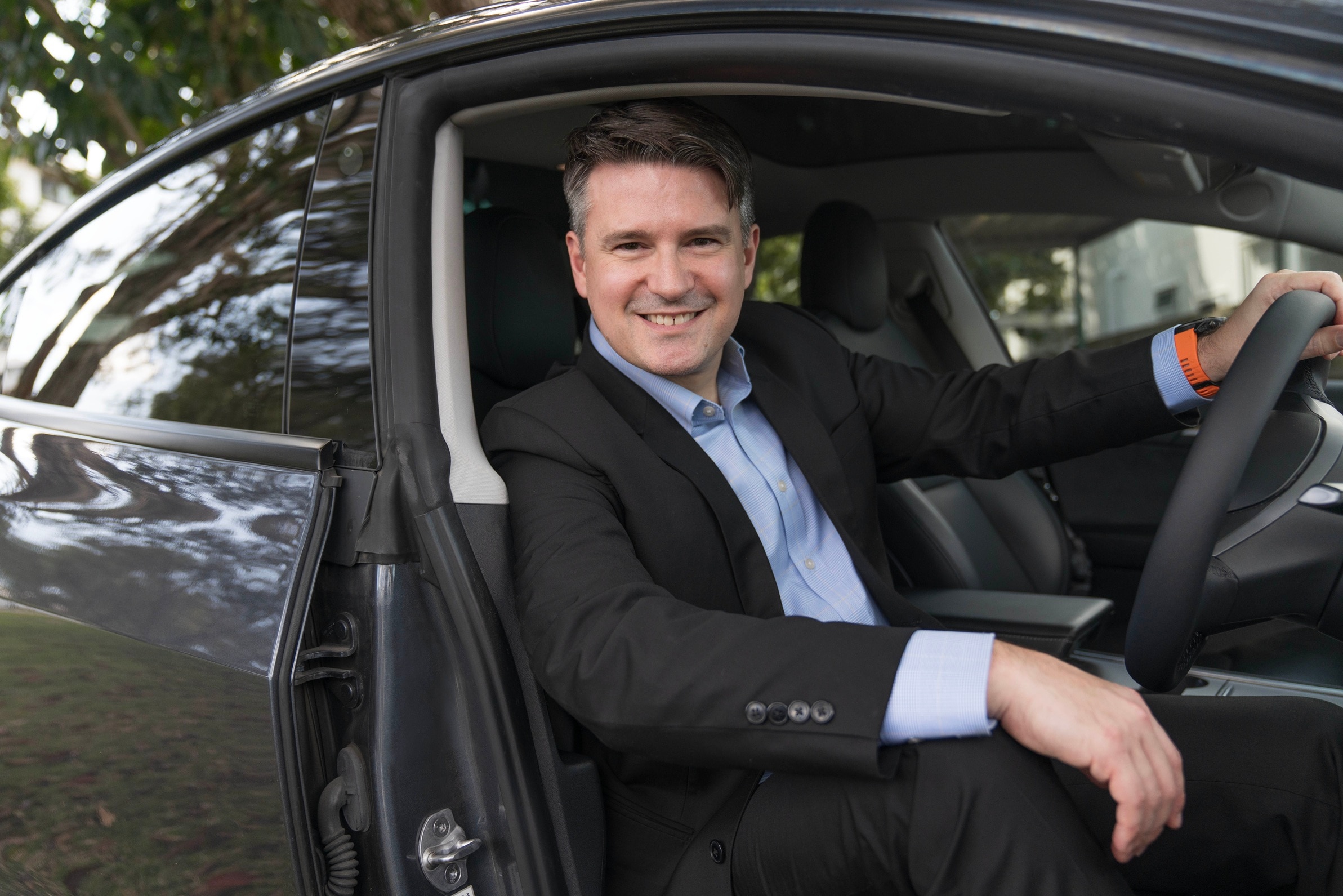 A smiling man in black suit and blue collared shirt sits in the drivers seat of an electric vehicle with the door open.