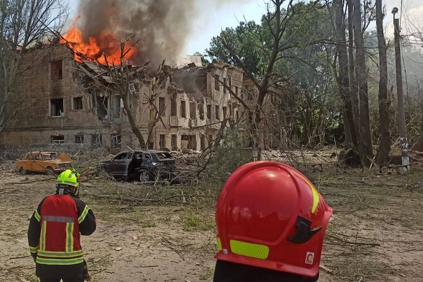 A building is caved in and on fire. Two rescue workers watch on. 