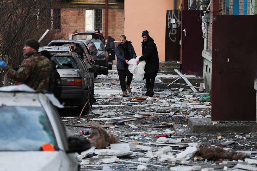 居民们走在满是废墟的街道上，两旁停满了损坏的汽车。