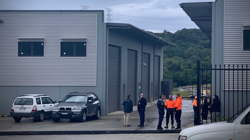 Police and workers in hi-vis vests stand outside an industrial site and talk to eachother