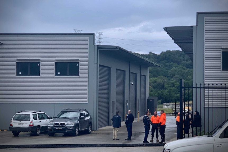 Police and workers in hi-vis vests stand outside an industrial site and talk to eachother