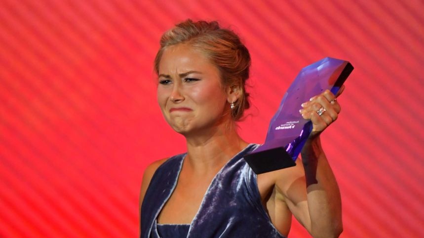 A young lady in a dark grey velvet dress holds up her Australian of the Year award.