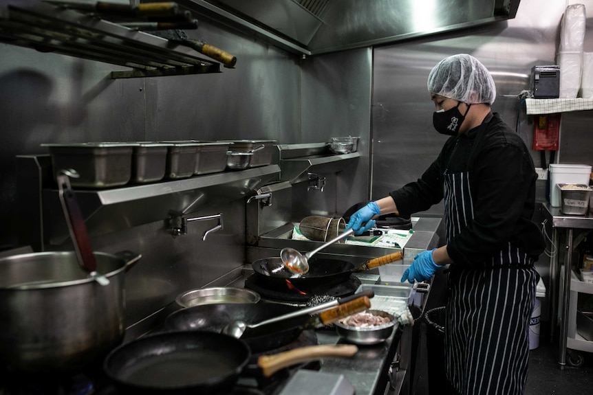 A man wearing a face mask stirs a large saucepan of food on a stove.