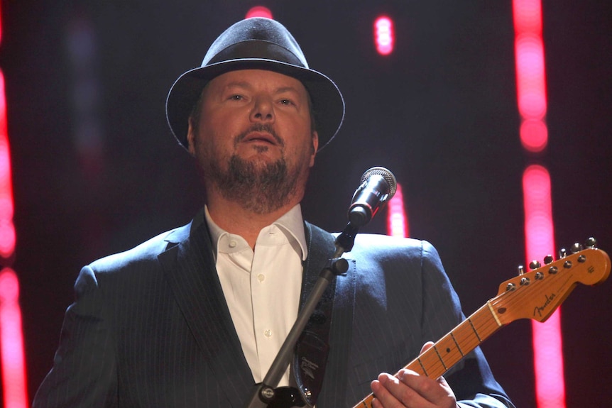 A white man in a suit and a hat plays a guitar