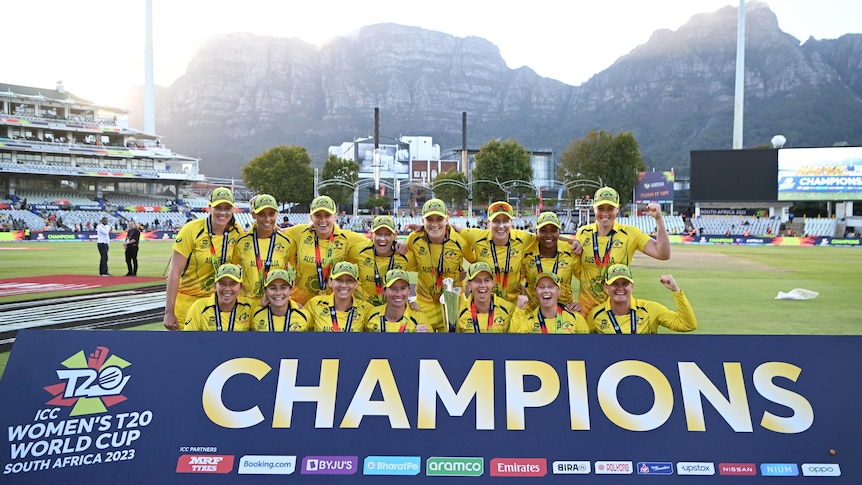 The Australian women's cricket team stands and crouches in front of a 'Champions' sign, with Table Mountain in the background.