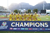 The Australian women's cricket team stands and crouches in front of a 'Champions' sign, with Table Mountain in the background.