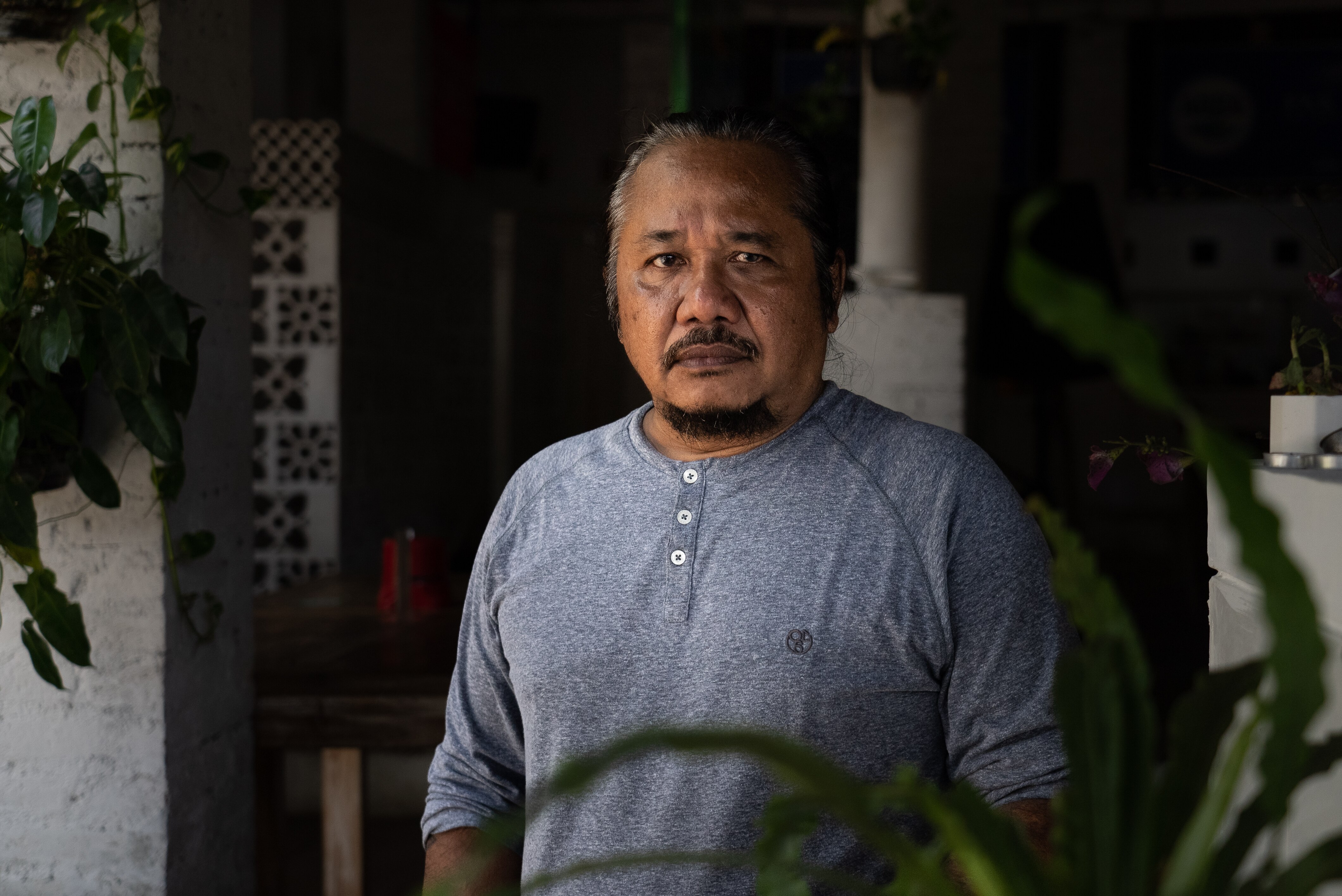A close up of a man with a beard and grey hair frowning near some pot plants.