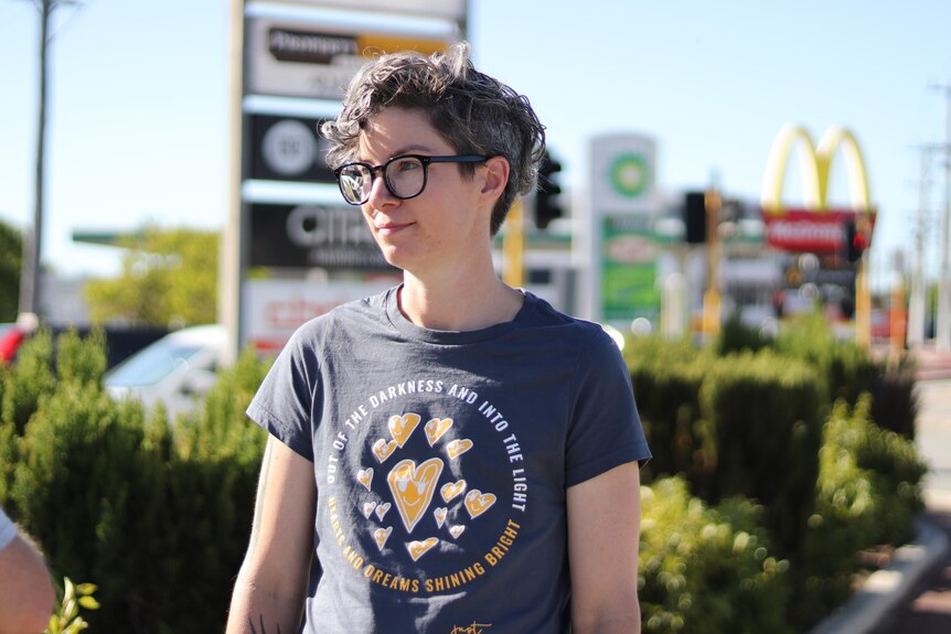 A young woman in a t-shirt and glasses stands in front of a busy intersection. 