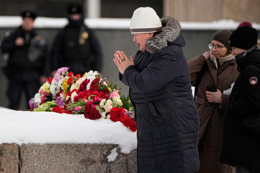 Police watch as a woman prays over a large bunch of flowers sitting on thick snow.