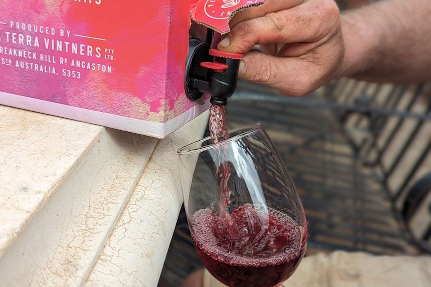 A hand pulls a tap in a box to pour out red wine into a glass.