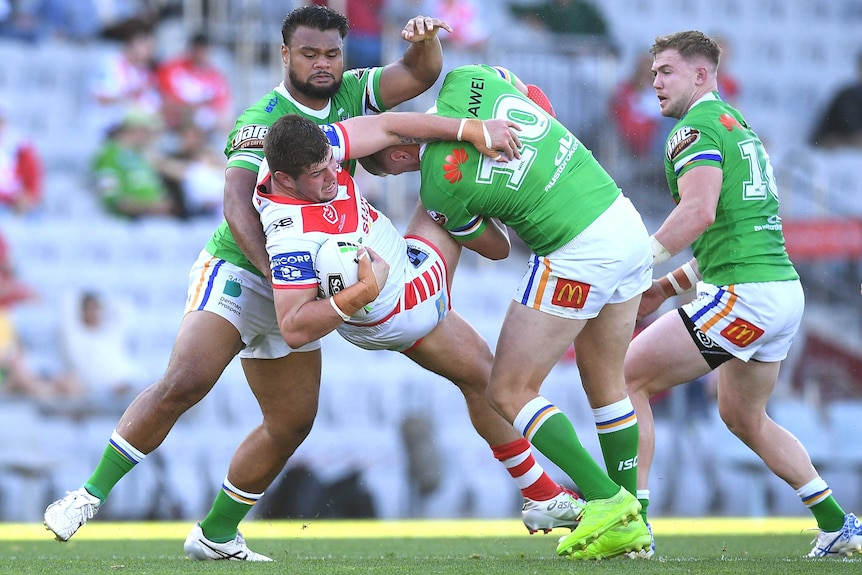 An NRL player is driven backwards in a tackle from two opposition players.