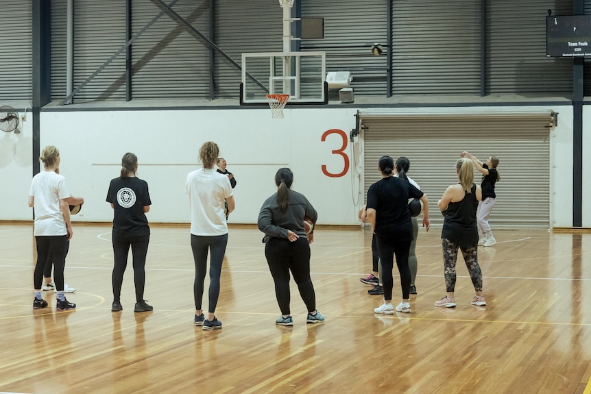 A photo from behind a group of women standing on a basketball court, listening to instructions from a coach.