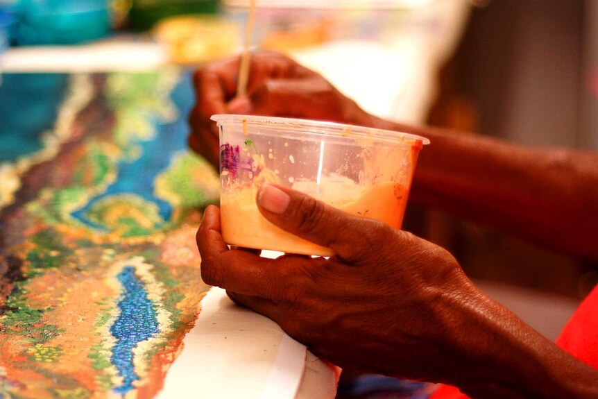 A close up shot of elder Ellery Sandy's hands as she paints on a colourful canvas.