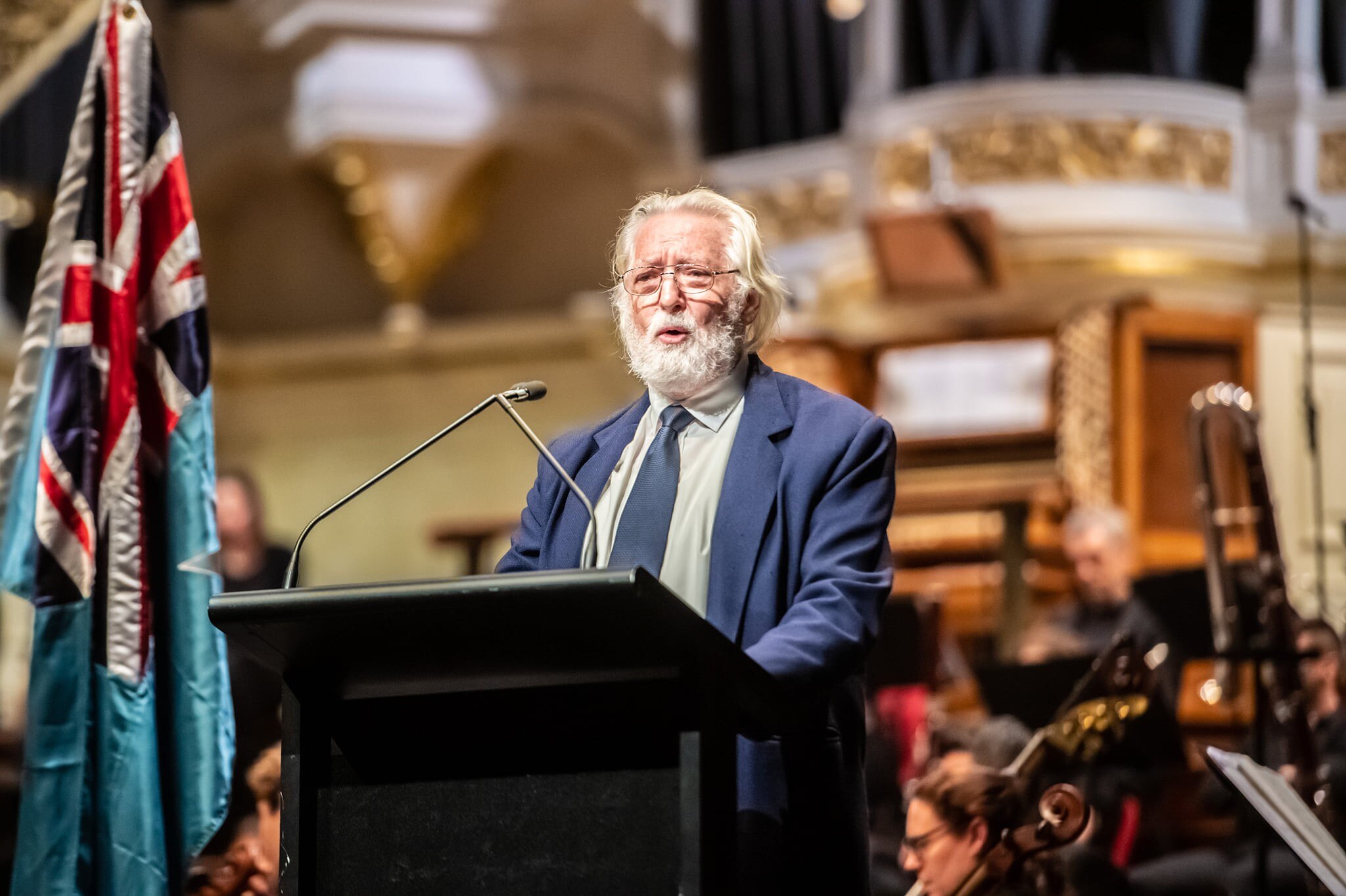 A man with white hair and beard stands at a lectern with an Australian flag, wearing a blue suit and tie